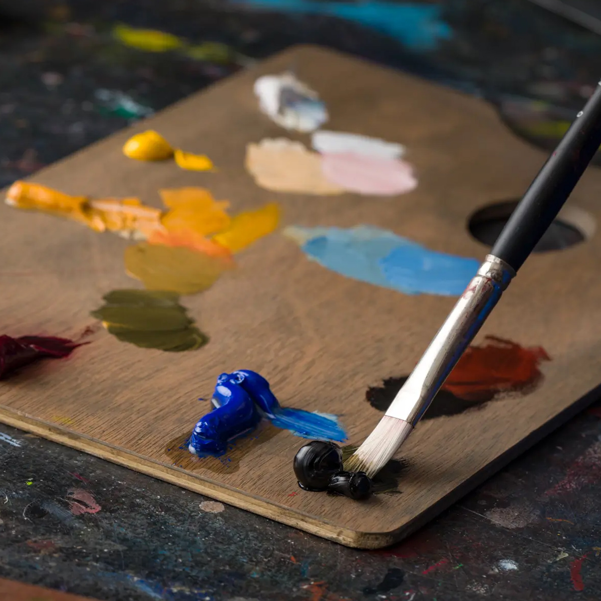 A paintbrush mixing black paint on a wooden palette with various colours