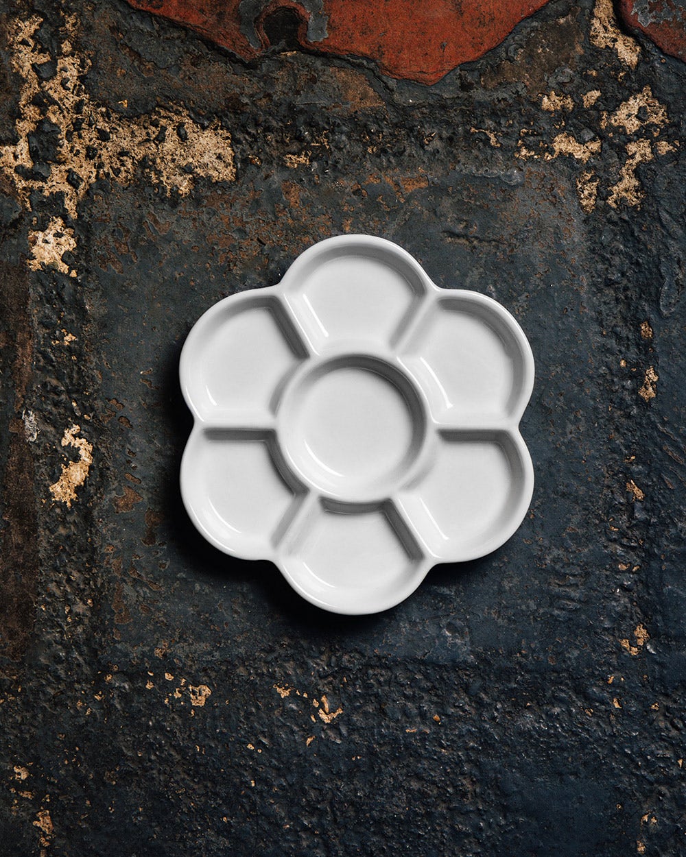 White ceramic palette in the shape of a flower, placed on a textured, rustic dark surface with earthy tones and weathered details.