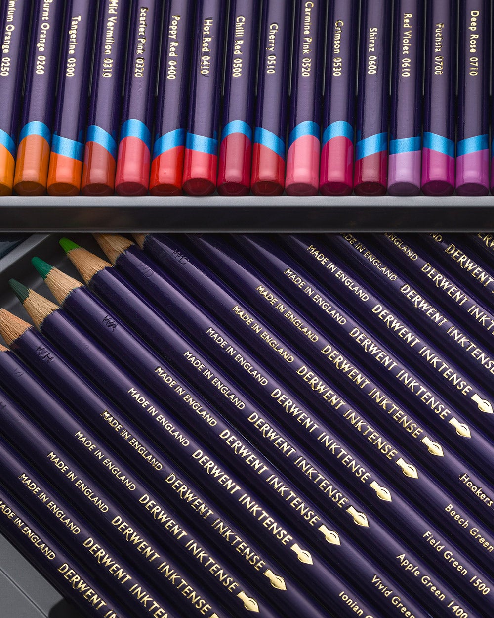 A set of Derwent Inktense colored pencils arranged in two layers within a metal tin. The top layer shows the pencil tips coated in vibrant shades of red, pink, orange, and purple.