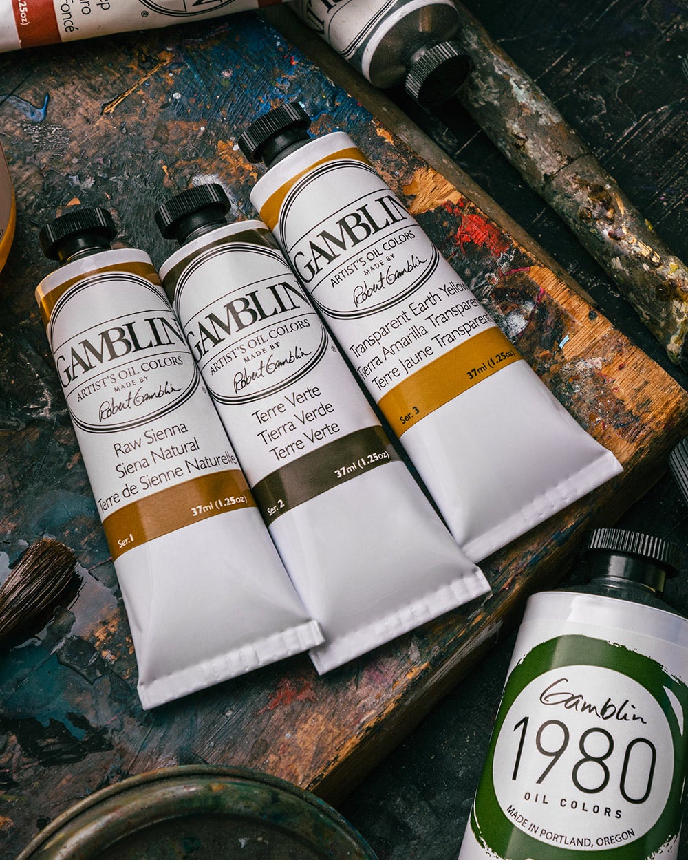 Three tubes of Gamblin artist's oil colors lying on a colorful, paint-splattered wooden palette. The tubes are labeled "Raw Sienna," "Terre Verte," and "Transparent Earth Yellow."