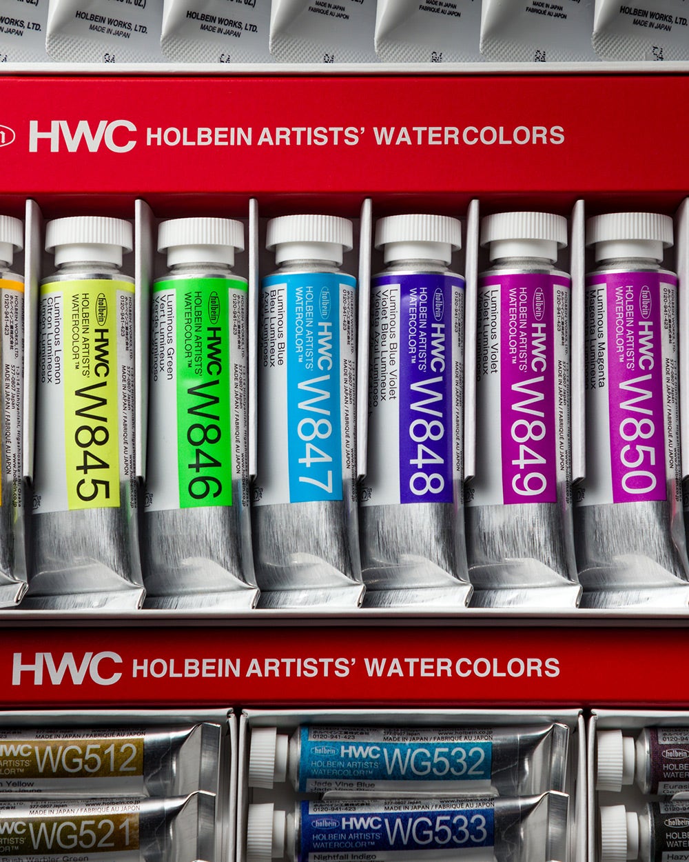 A close-up photograph of a set of Holbein Artists' Watercolors in metal tubes, neatly arranged in a red box. The tubes are labeled with color names and codes, such as W845 (Luminous Yellow), W846 (Luminous Green), W847 (Luminous Blue).