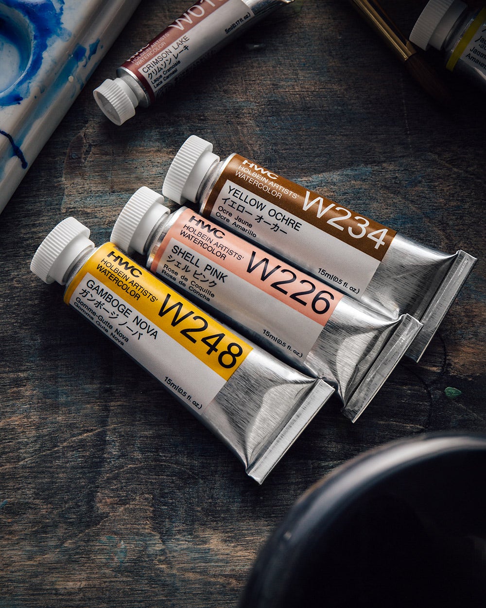 Close-up of Holbein Artists' Watercolor tubes in Yellow Ochre, Shell Pink, and Gamboge Nova, arranged on a textured wooden surface with art supplies in the background.