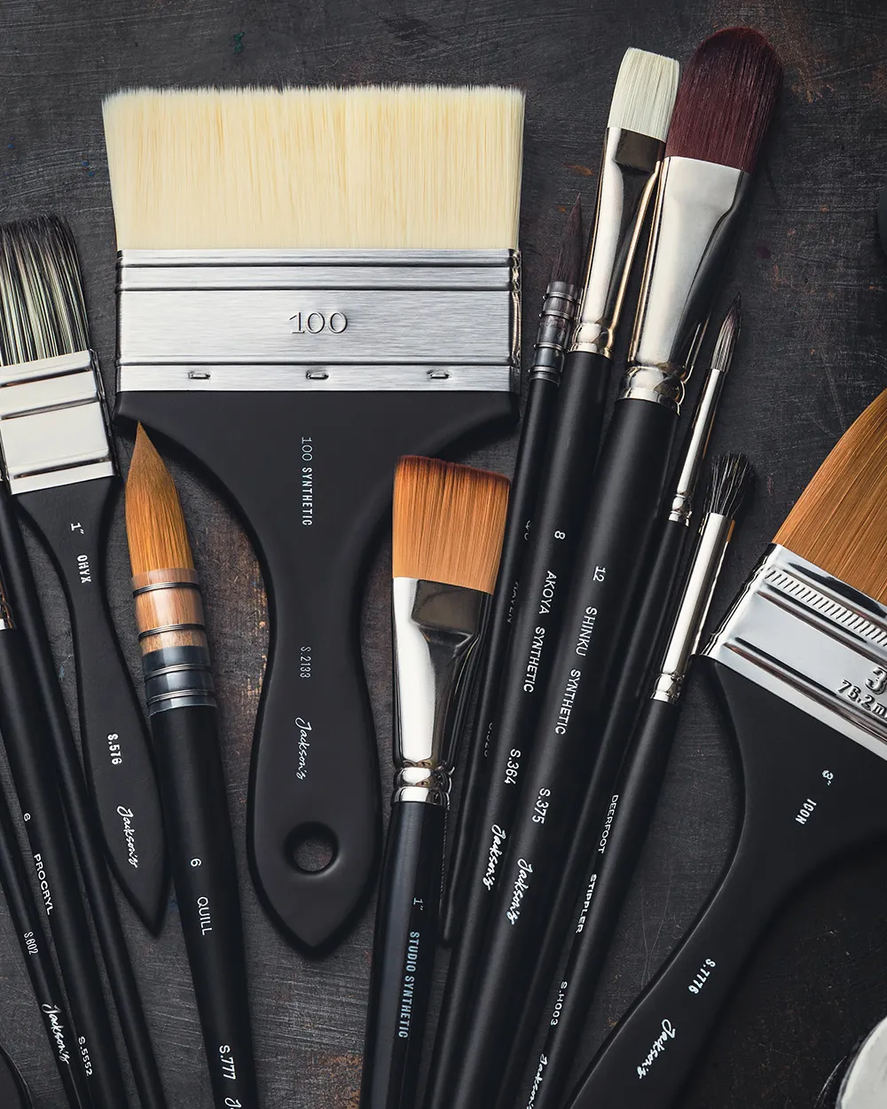 A variety of artist paintbrushes laid out on a dark surface. The brushes have sleek black handles with white text indicating types and sizes, including wide flat brushes, round brushes, and angled brushes. 