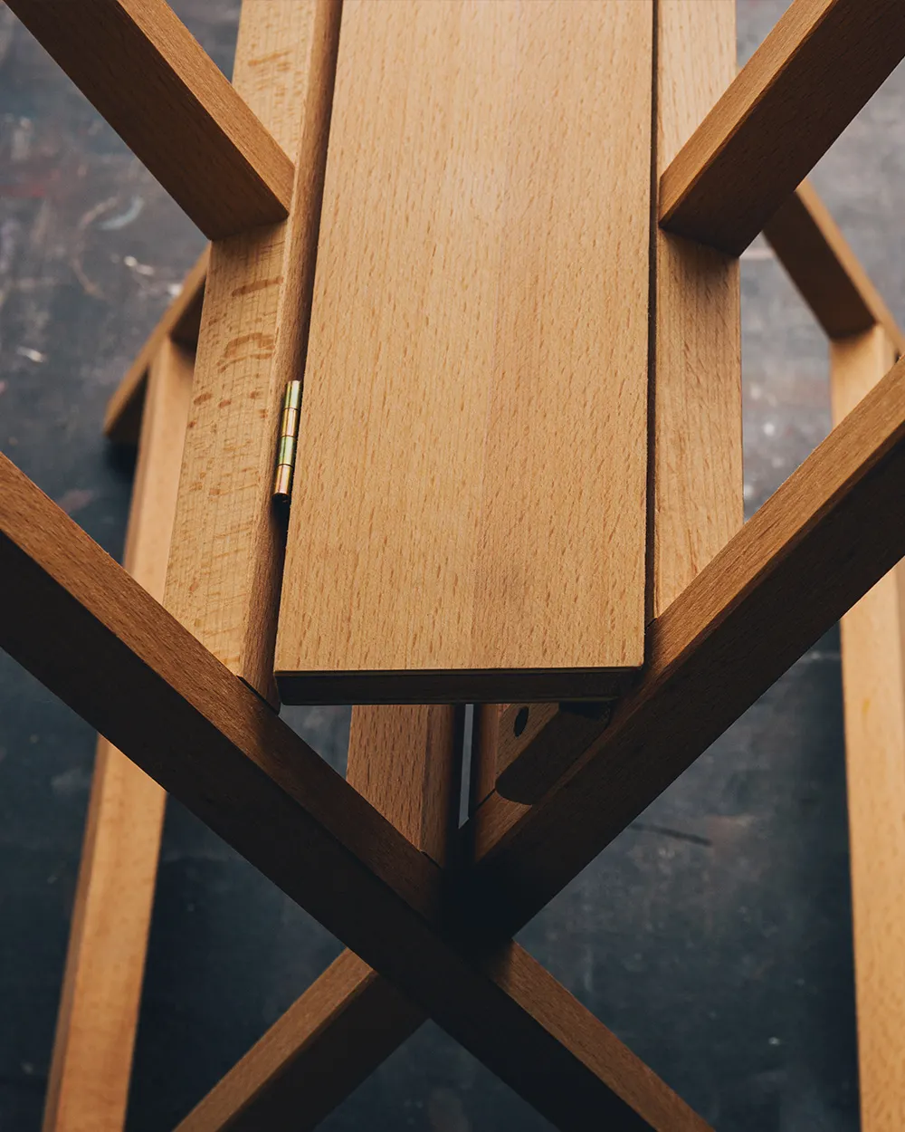  Close-up view of a wooden folding structure, likely a print rack or artist's drying rack, with a light natural wood finish. The image shows part of the hinge mechanism and intersecting wooden legs on a dark speckled surface.