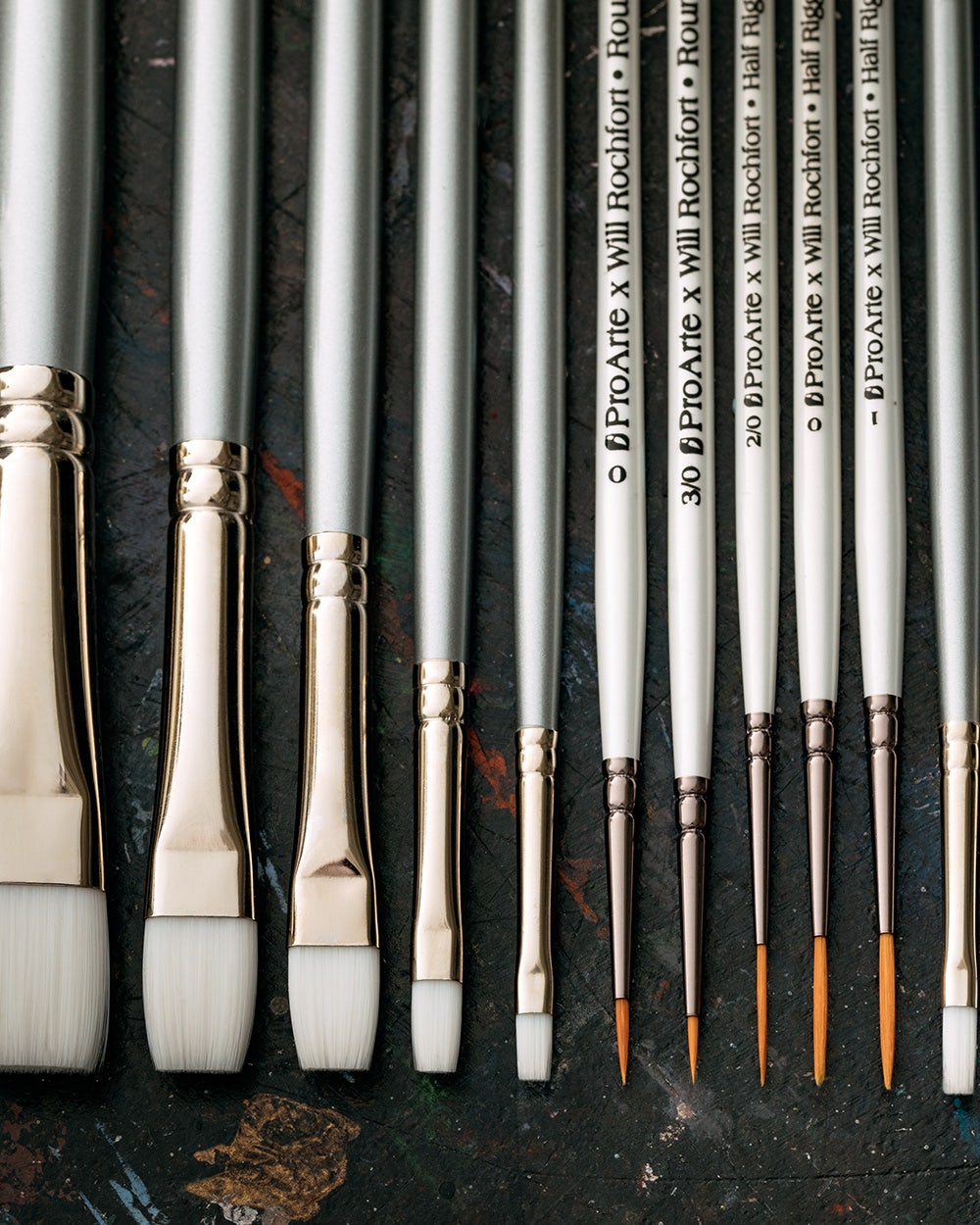 A neatly arranged set of twelve artist paintbrushes with silver handles and white or brown bristles, displayed on a dark, paint-splattered surface. The brushes vary in size and shape, including flat, filbert, and round tips.