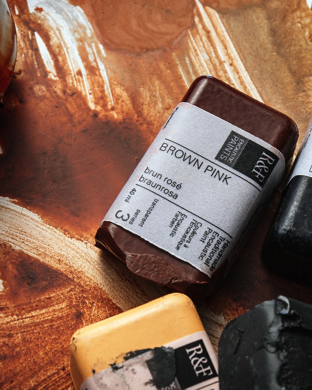 "A close-up photograph of several R&F Handmade Paints encaustic paint blocks resting on a textured, paint-stained surface. The blocks include colors such as Brown Pink and Payne’s Grey, with their labels partially visible.