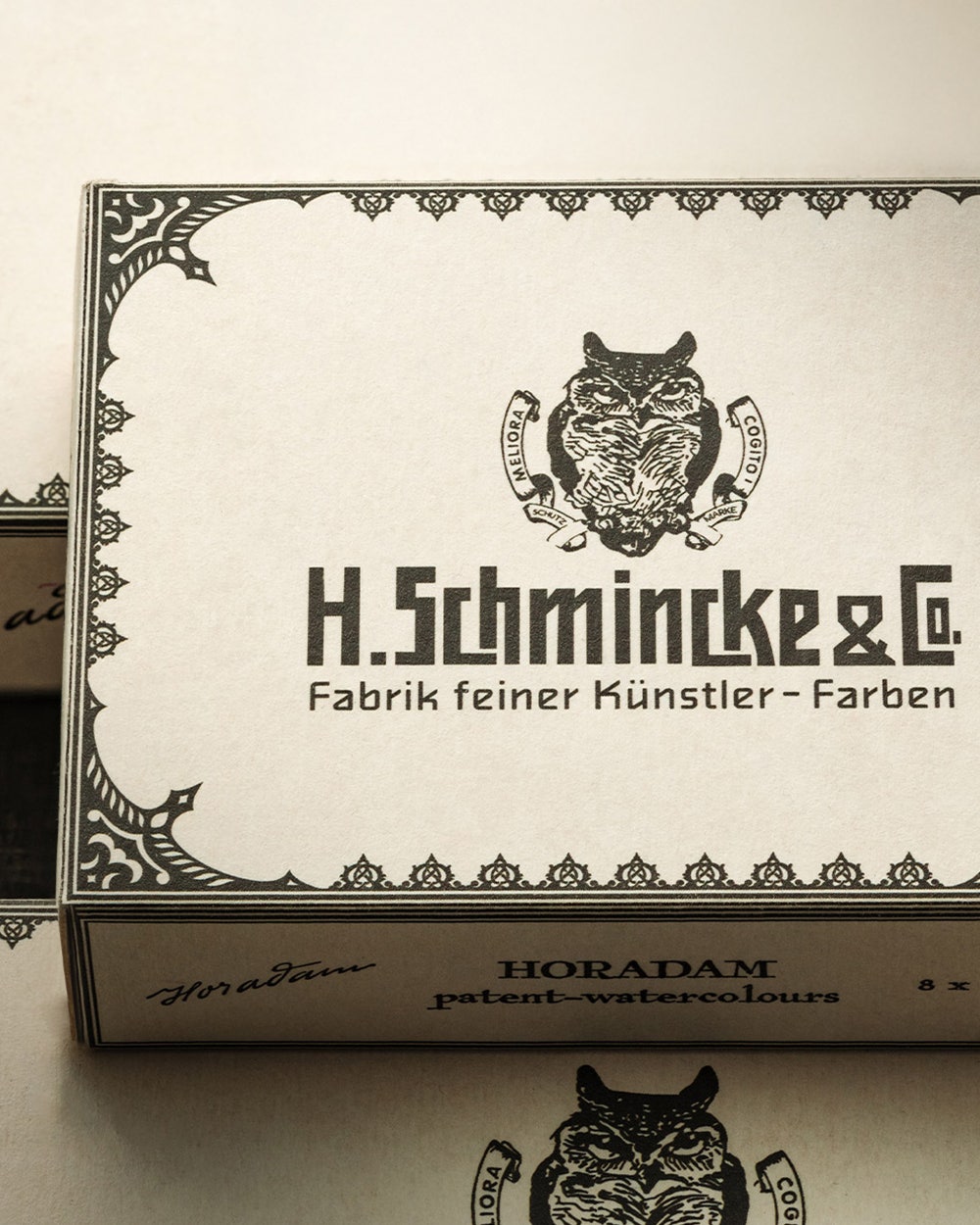 Vintage-style packaging of H. Schmincke & Co. artist paints, featuring intricate decorative borders, an owl emblem with Latin phrases, and bold, old-fashioned typography. The text highlights the brand’s heritage and product quality.
