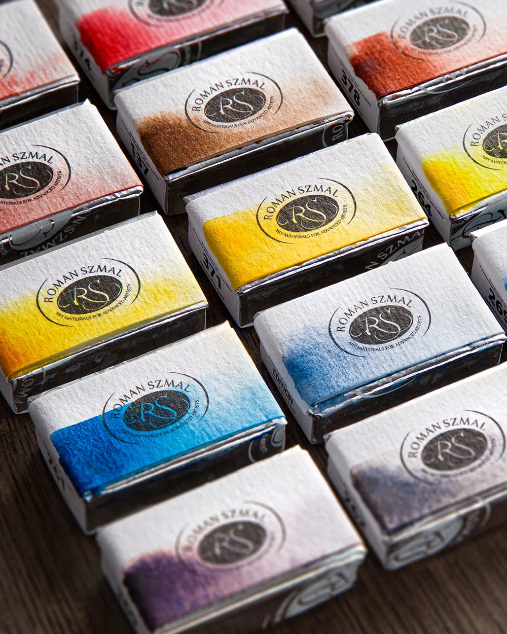 A close-up view of several watercolor paint pans from the brand Roman Szmal, neatly arranged on a wooden surface. Each pan is wrapped in a white label with the brand's logo and a color swatch indicating the paint inside.