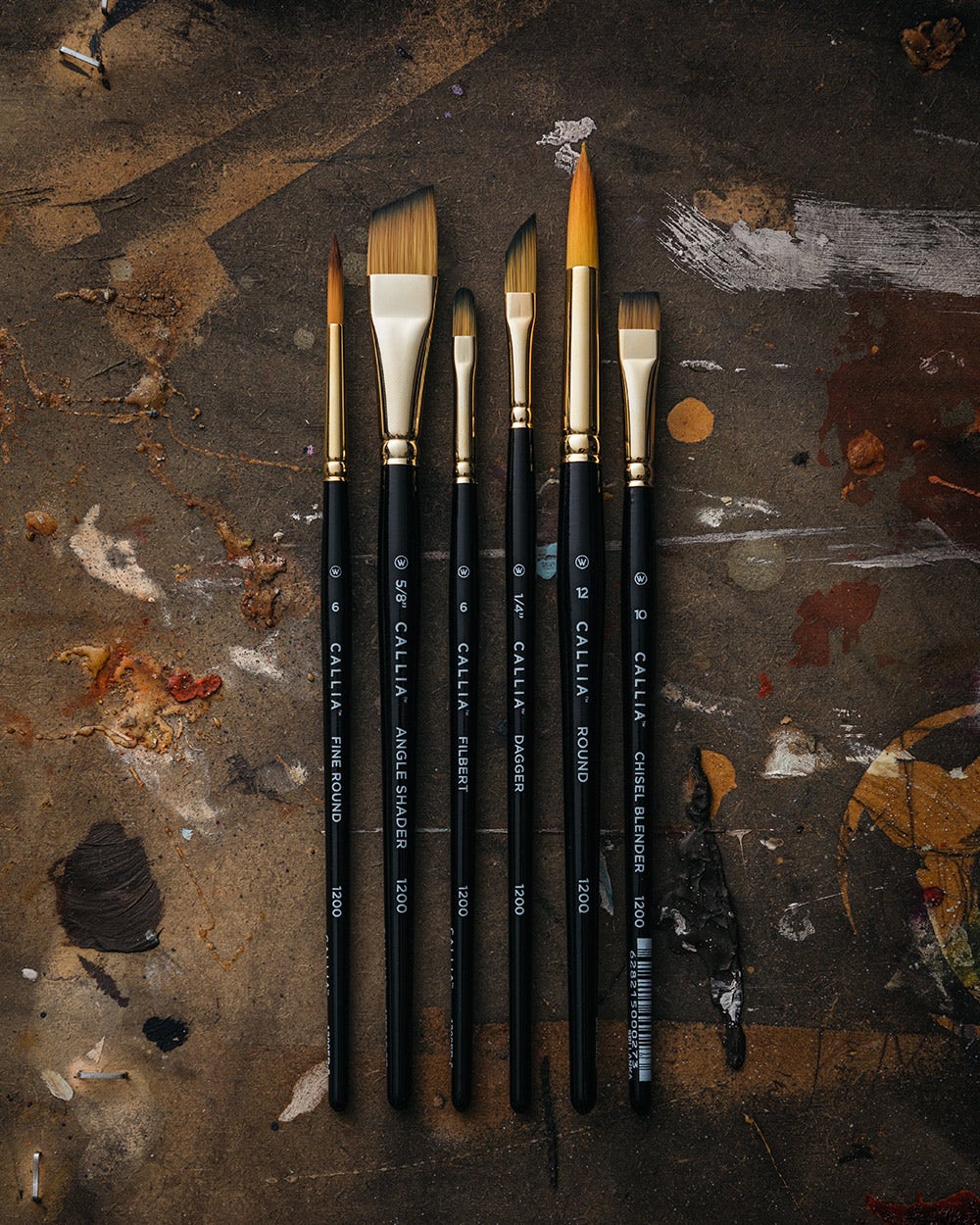 A set of six Callia paintbrushes arranged in a row on a paint-splattered surface. The brushes vary in size and shape, including a fine round, angle shader, filbert, dagger, round, and chisel blender.