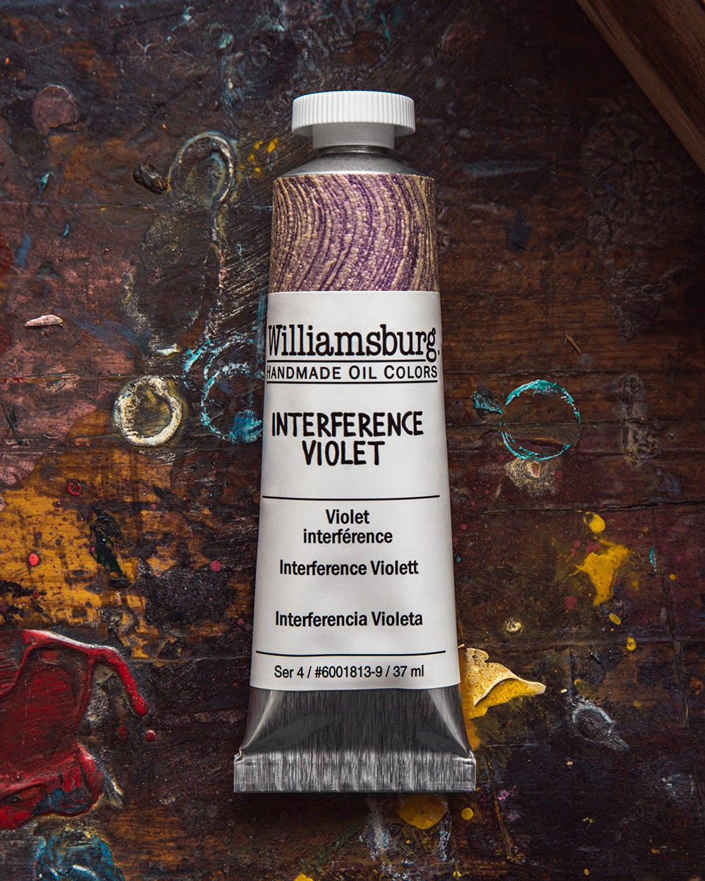 A tube of Williamsburg Handmade Oil Colors in the shade "Interference Violet" is placed on a well-used, colorful artist's work surface. The label on the tube features the paint name in English, French, German, and Spanish