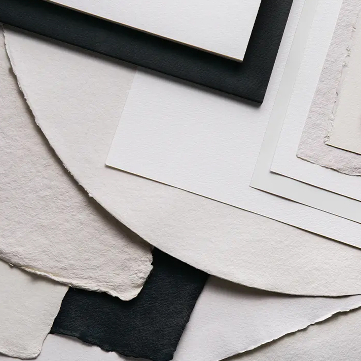 An arrangement of various types of paper, including textured, smooth, and deckle-edged sheets, layered in different shapes and shades of white, cream, and black. The composition highlights the diverse textures and finishes of the papers.
