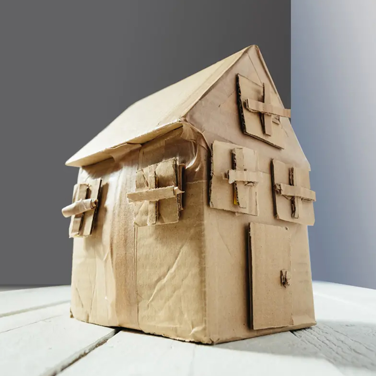 A small cardboard house model with a pitched roof, shutters on the windows, and a door, all crafted from brown cardboard.