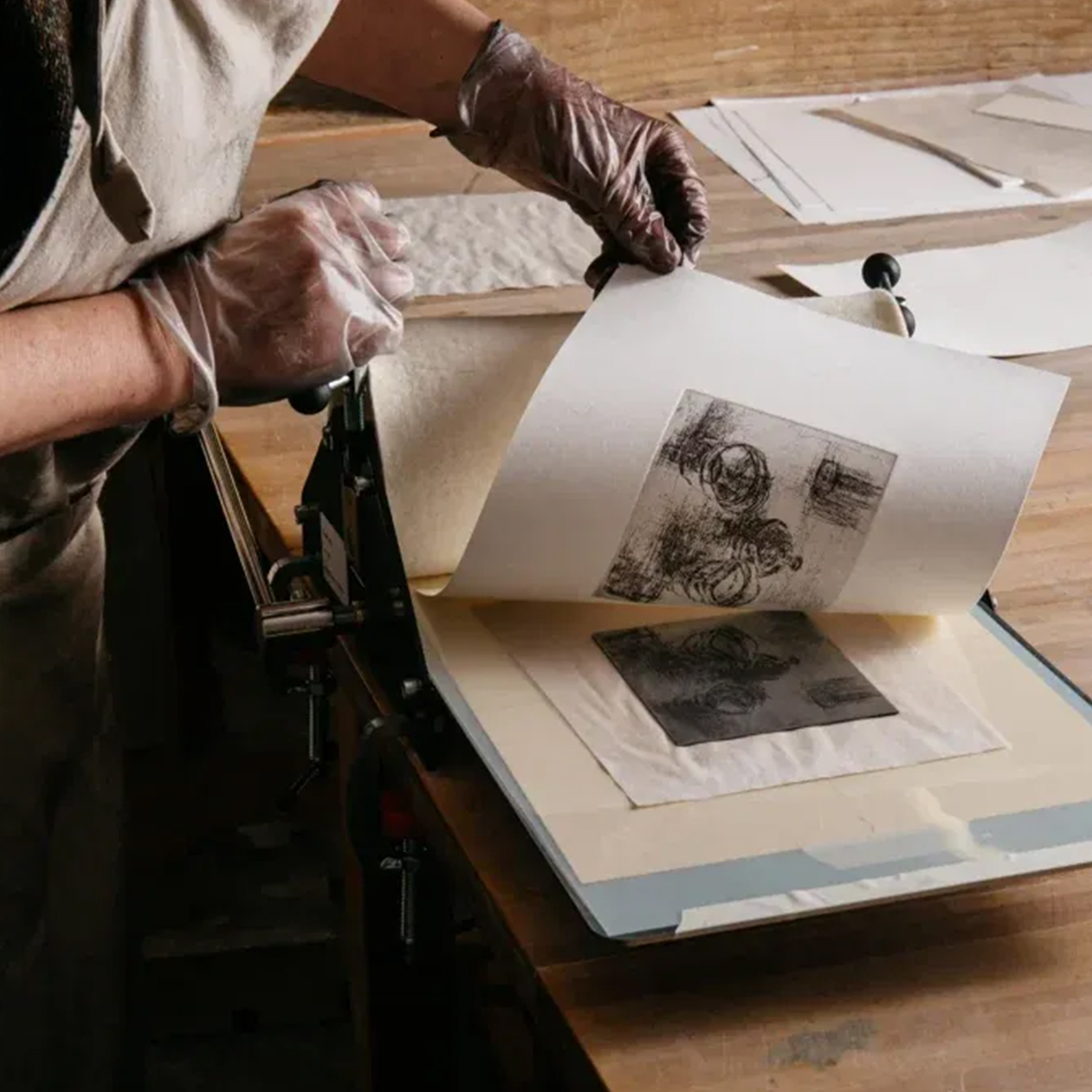 A person wearing disposable gloves is lifting a printed sheet from an etching press during the intaglio printing process. The print features detailed black ink artwork, and the plate is visible beneath the paper. The workspace includes a wooden table with