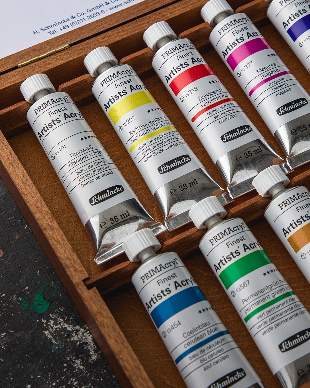 A wooden box containing several tubes of Schmincke PRIMAcryl Finest Artists' Acrylic paint in various colors, including titanium white, cadmium yellow light, vermilion red, magenta, cerulean blue, and permanent green. 