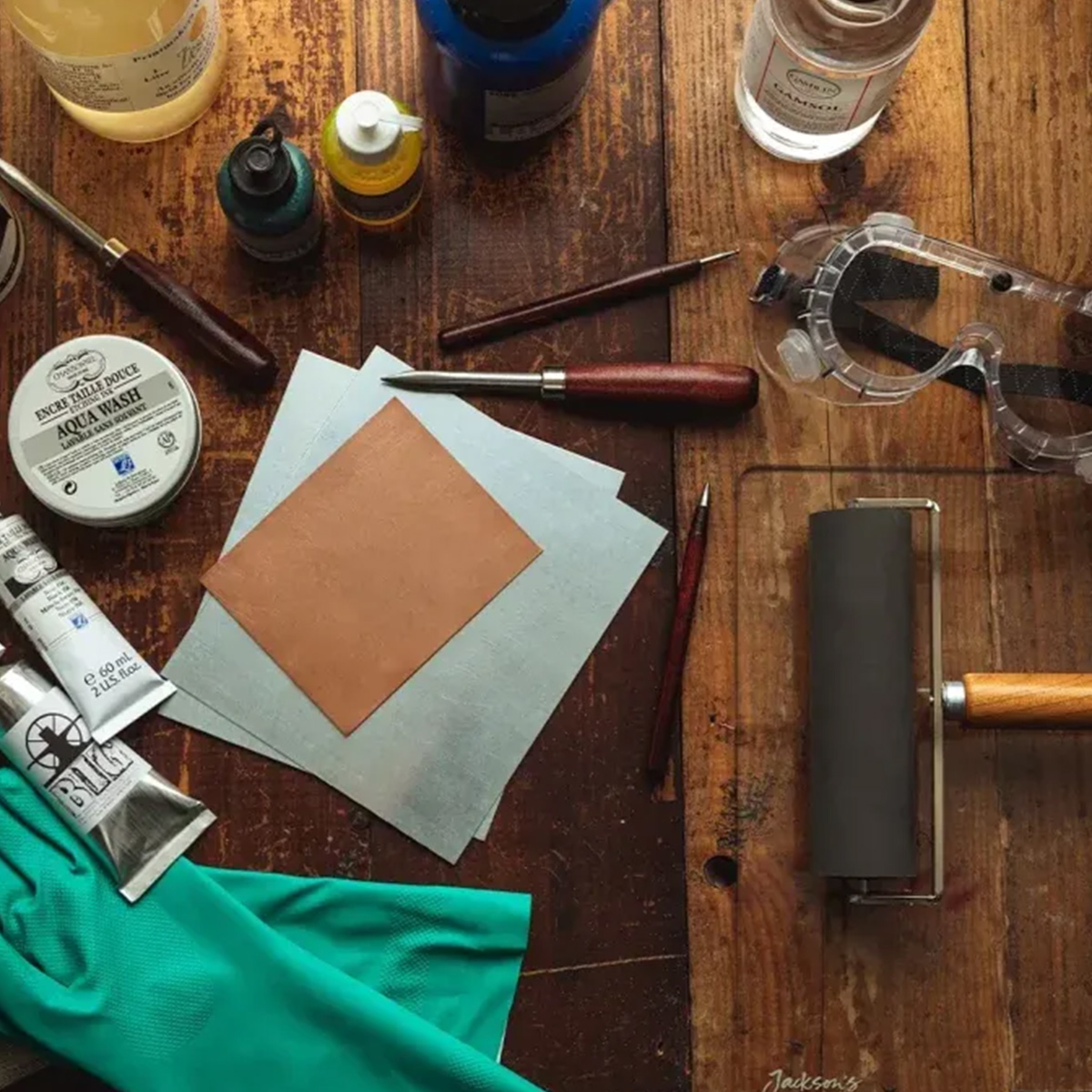 A flat lay of tools and materials for intaglio printmaking on a wooden table. Items include a copper plate, safety goggles, a rubber brayer, inks in various colors, a palette knife, a burnishing tool, a jar of Aqua Wash, tubes of ink, green gloves, and cl