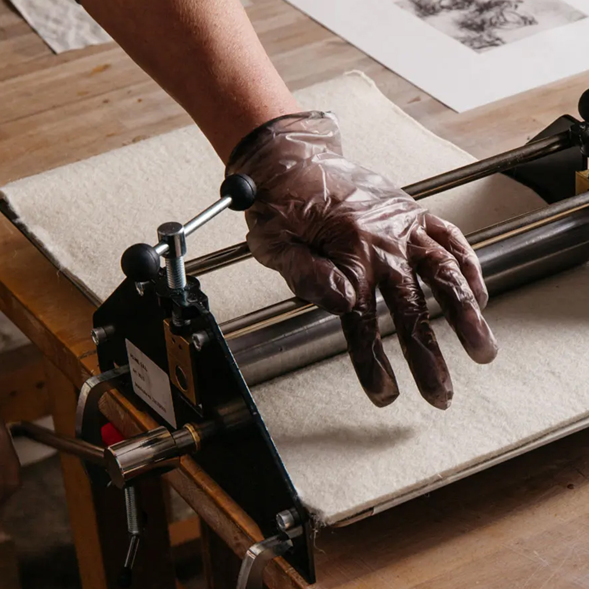 A close-up of a person’s hand wearing a disposable glove operating a small etching press on a wooden table. The press is set up with a felt blanket, and a print is visible in the background, suggesting an intaglio printmaking process in progress. The work